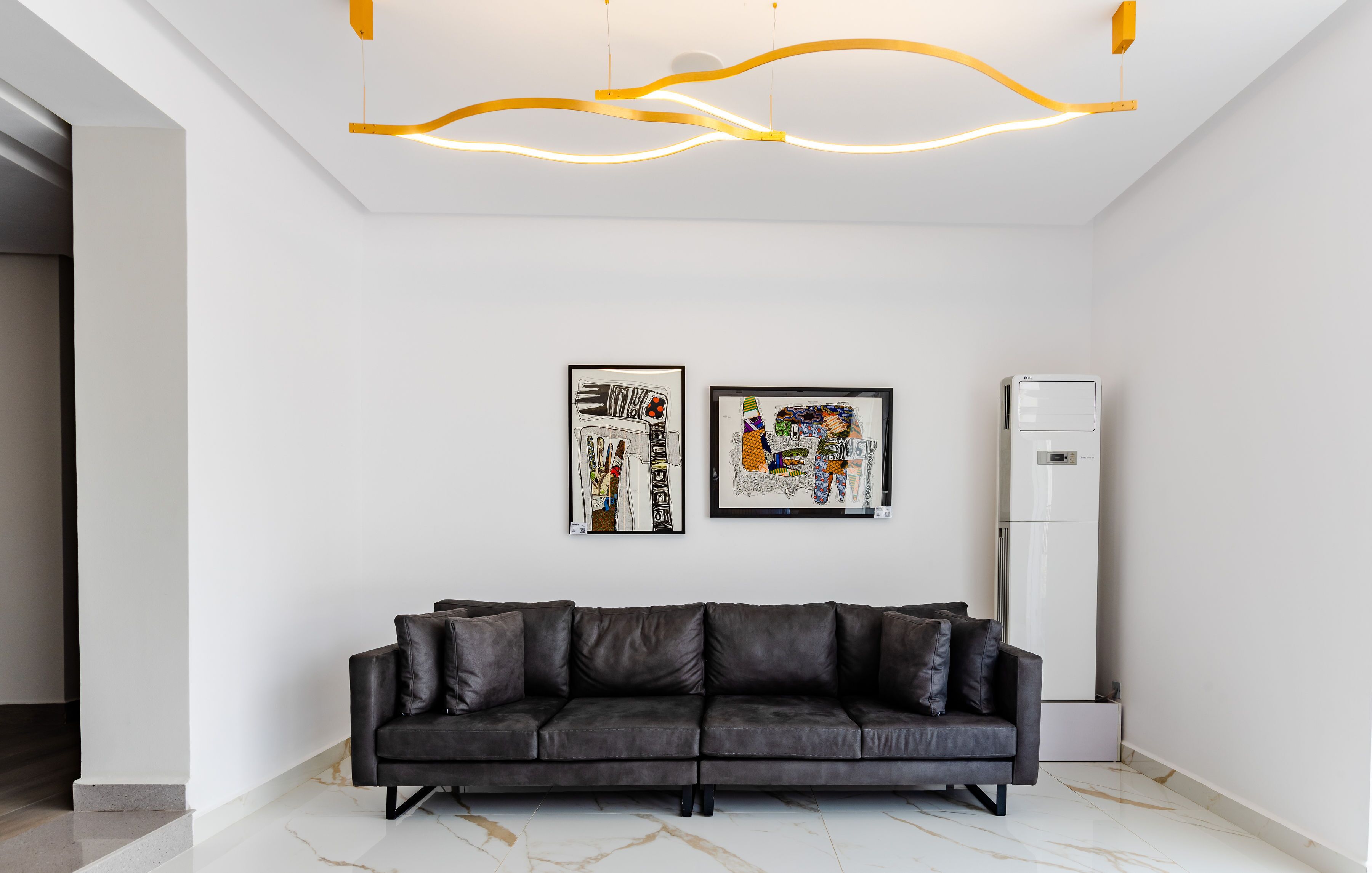 A modern living room with a black leather sofa, two framed artworks, and a white air conditioning unit.