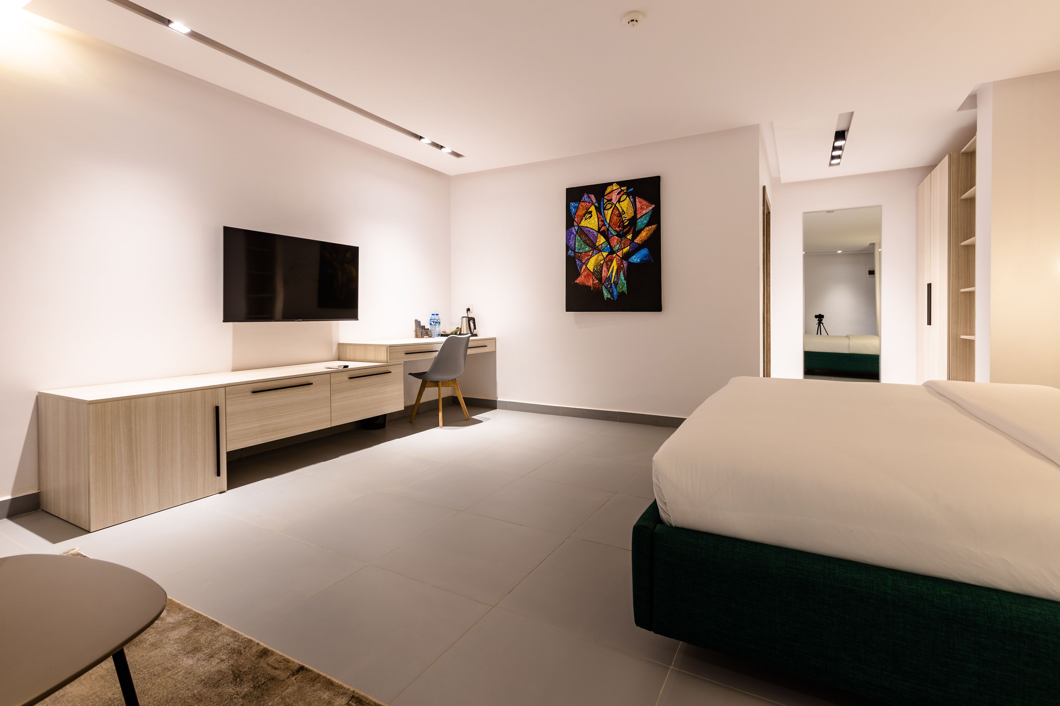A modern bedroom with a bed, a desk, a chair, a TV, and a colorful artwork on the wall.
