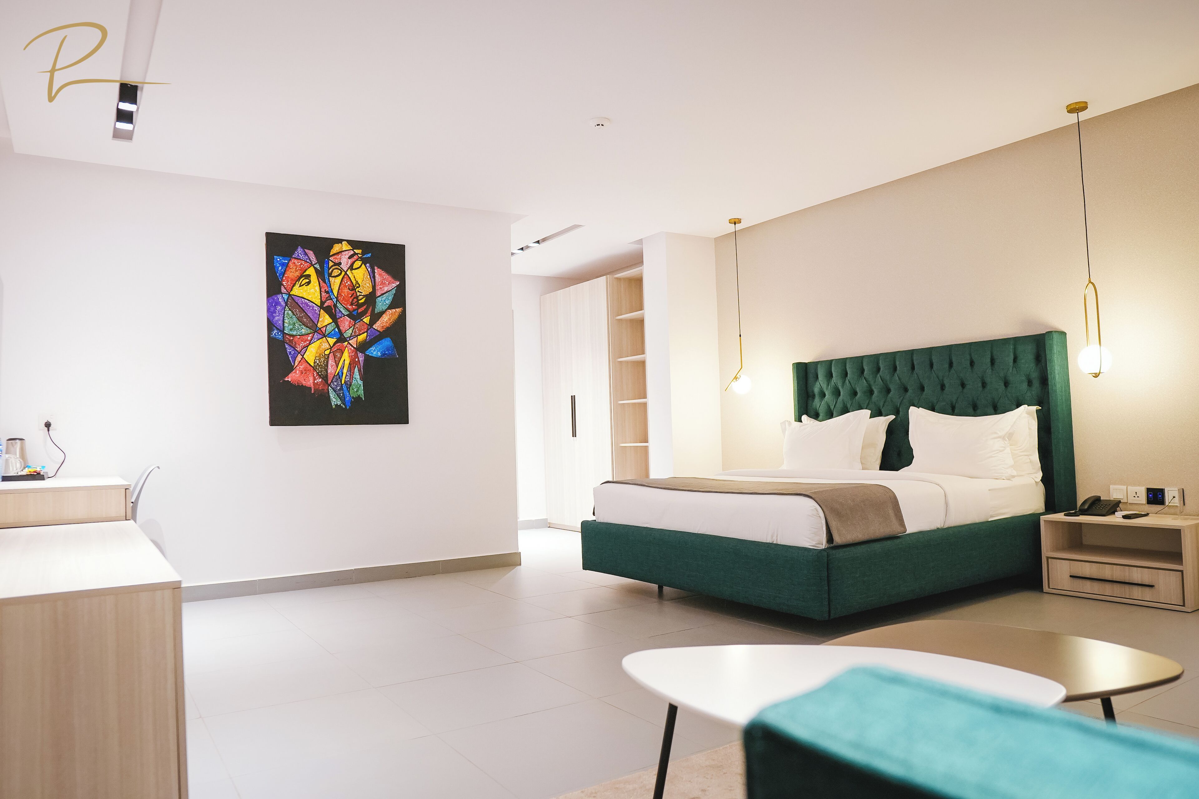 A modern hotel room with a green upholstered bed, a desk with a chair, a bedside table, and a wall-mounted artwork.