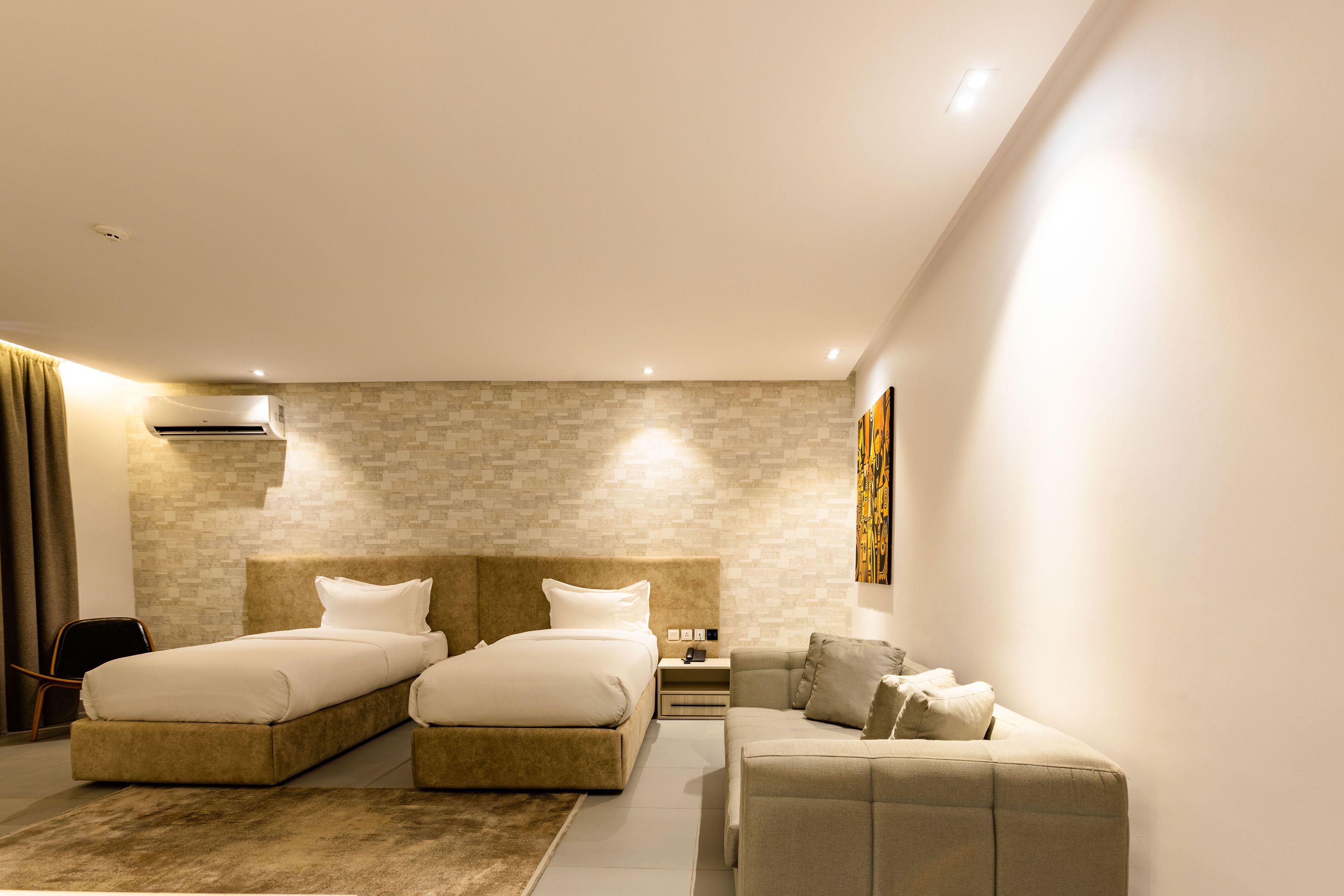 A modern hotel room with two beds, a sofa, a nightstand, and a wall-mounted air conditioner.