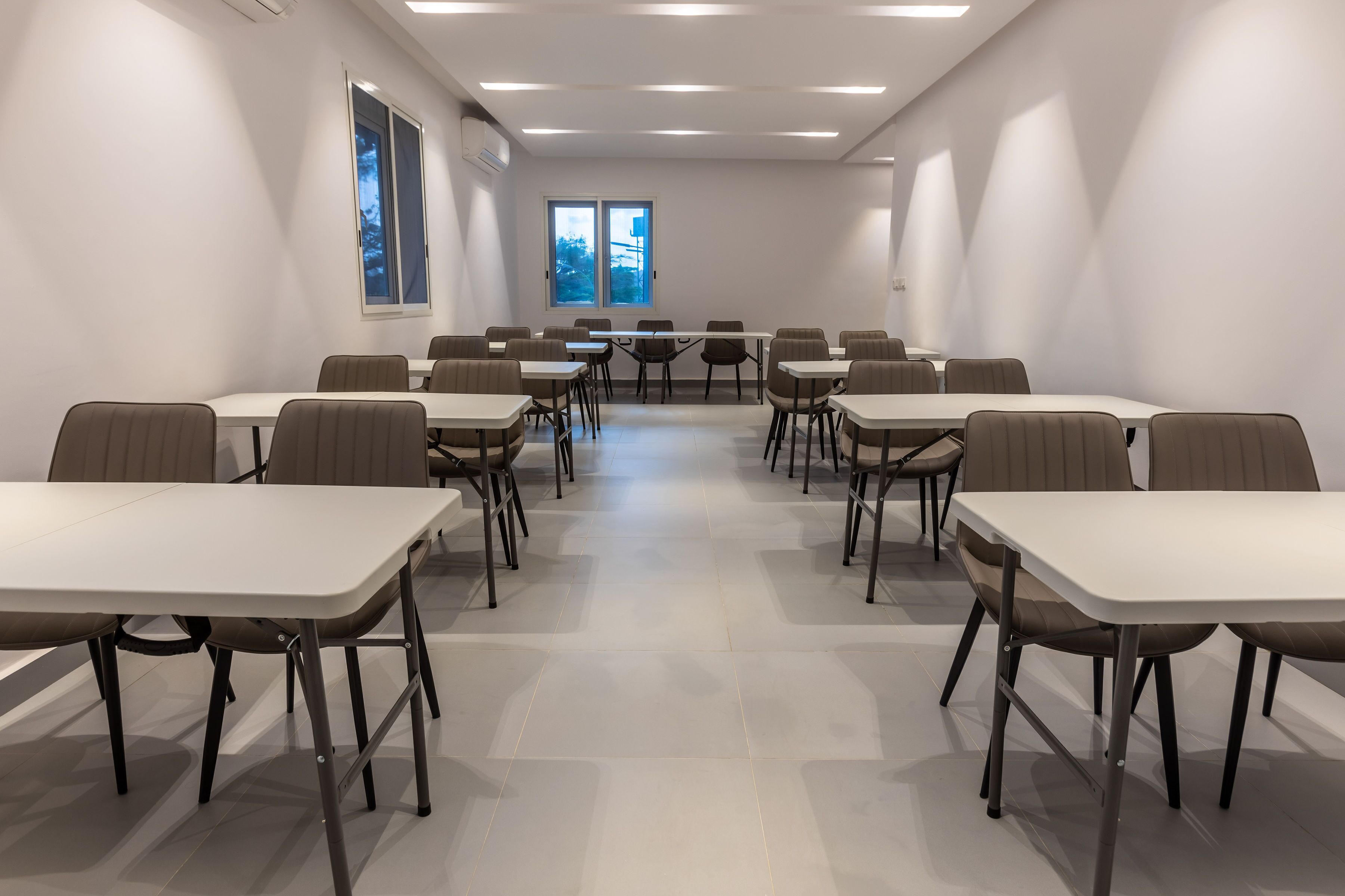 Un salón de conferencias con mesas y sillas blancas, ventanas amplias y vistas a árboles.