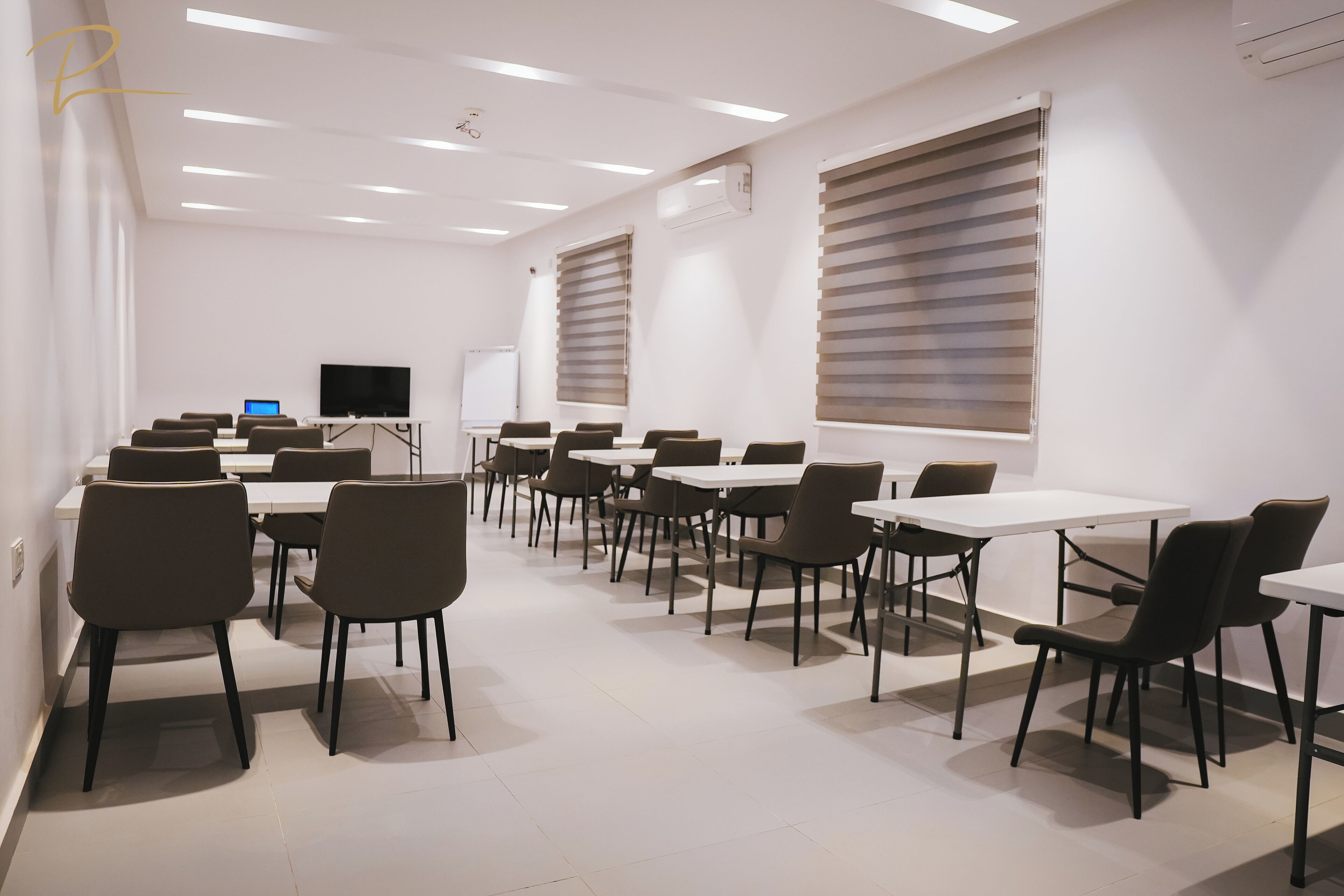 A conference room with white walls, multiple tables and chairs, a large screen, and blinds on the windows.