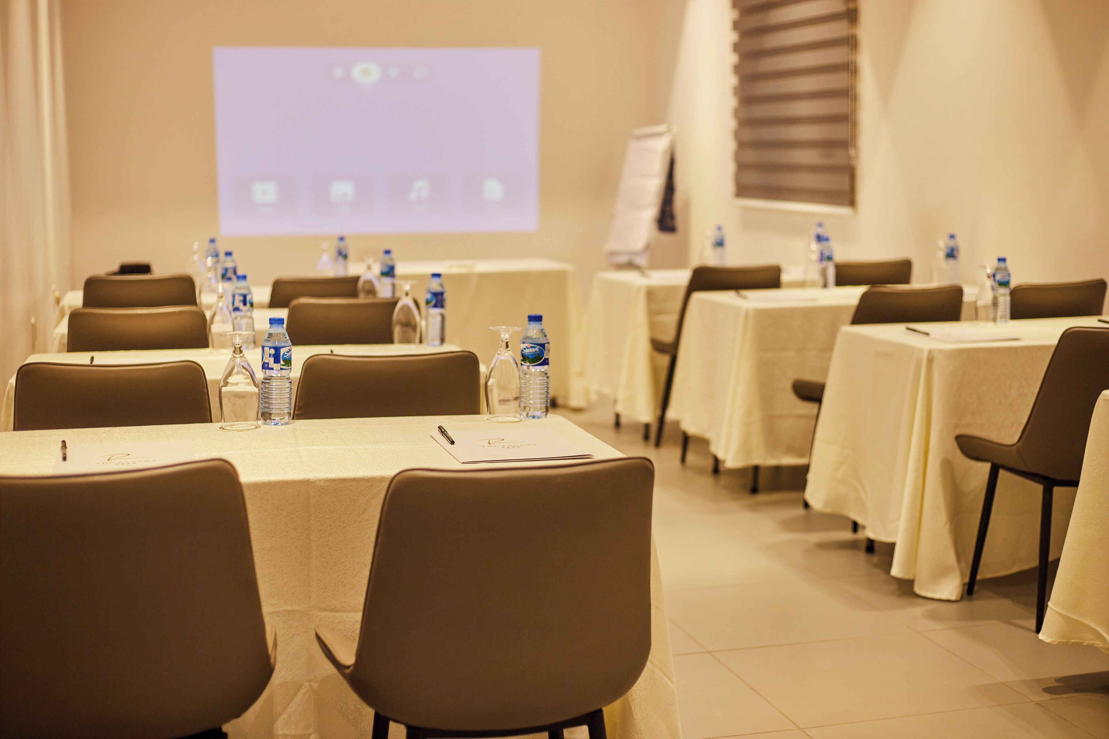 Une salle de conférence équipée de tables, de chaises, de bouteilles d’eau et d’un écran de présentation.