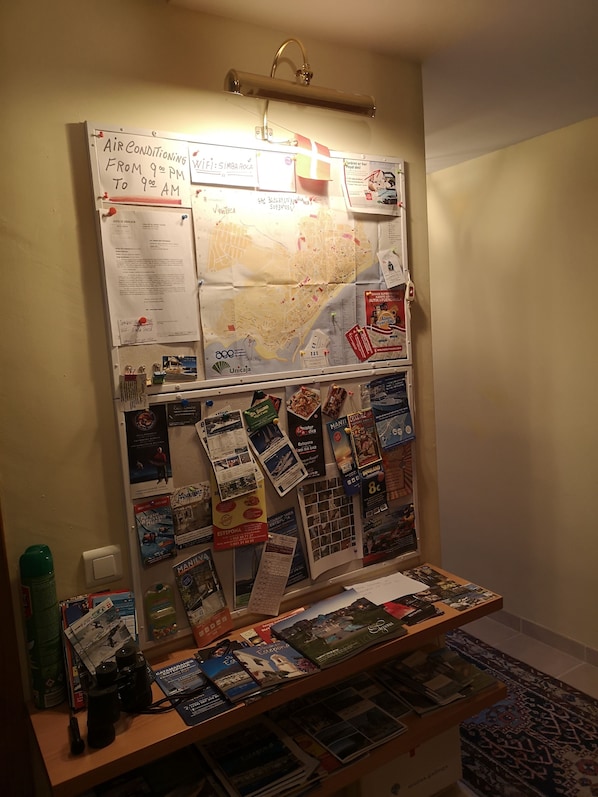 Un panneau d’affichage où sont accrochées diverses brochures de voyage, une carte et un panneau indiquant les heures de fonctionnement de la climatisation.