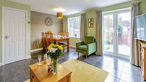 A dining area with a table set for four, a green armchair, a TV, and a large window with curtains. - 3 bedroom accommodation in Upper Soudley (Upper Soudley)