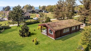 Une petite cabane en bois à un seul étage, avec un toit recouvert d’herbe, entourée d’une pelouse bien entretenue et d’arbres.