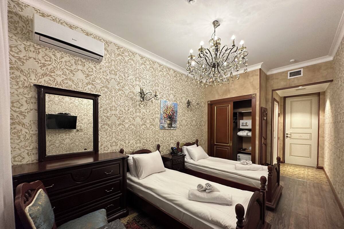 A hotel room with two beds, a large mirror, a chandelier, a wardrobe, and a television.