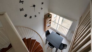 Una habitación con un reloj de pared grande, una escalera de caracol, una mesa de comedor con sillas y una ventana con vistas al exterior.