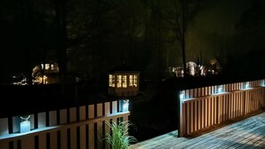 Une terrasse en bois avec une rambarde, un pot de fleurs et des structures illuminées en arrière-plan.