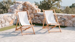 To træhytter med hvide sæder og ryglæn, placeret på en stenbelagt terrasse med havudsigt og en stenvæg i baggrunden.