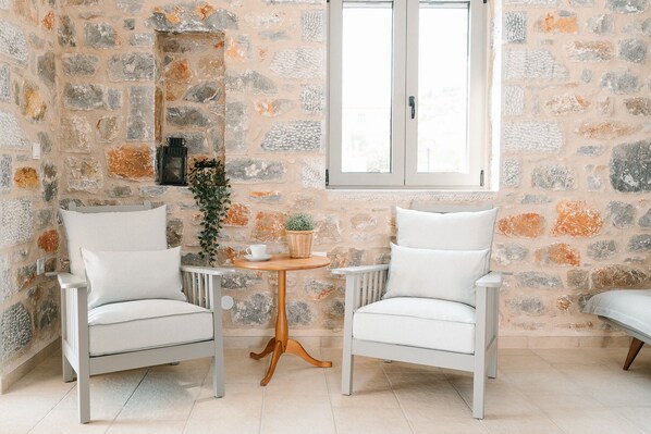 Une pièce avec deux fauteuils blancs, une petite table en bois sur laquelle se trouvent une tasse blanche et un panier, et un mur en pierre avec une fenêtre.