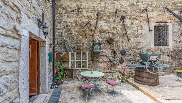Un patio con paredes de piedra, una puerta de madera, una mesa redonda con sillas y una carretilla.