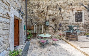 Une cour bordée de murs en pierre, avec une porte en bois, une table ronde entourée de chaises et une brouette.