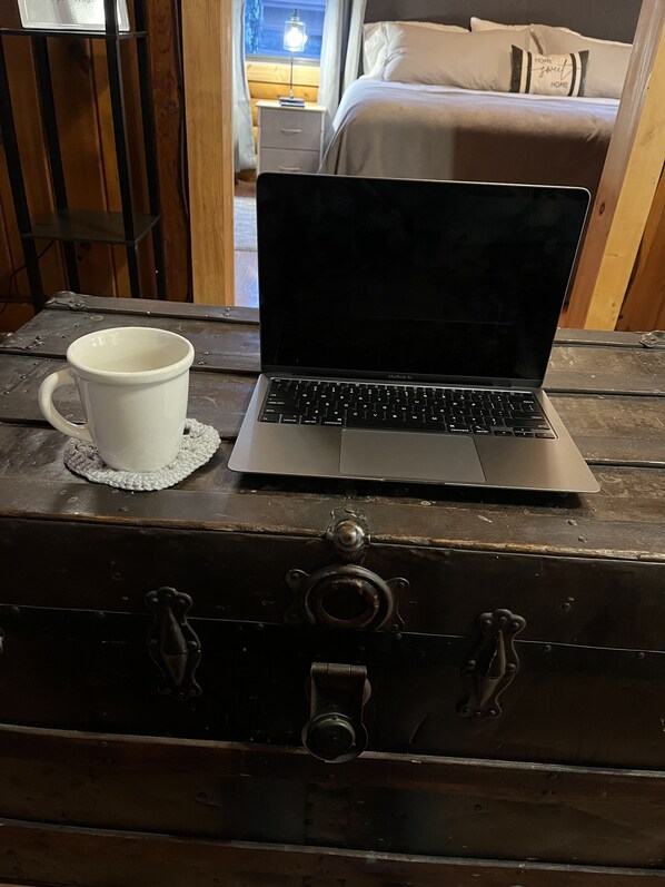 Ein Laptop auf einer hölzernen Kommode, eine weiße Tasse auf einem Untersetzer und ein Bett mit Kissen im Hintergrund.