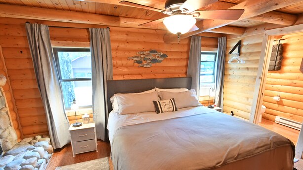Ein gemütliches Schlafzimmer in einer Blockhütte mit Bett, Nachttischen, Deckenventilator und einem Fenster mit Vorhängen.