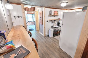 Eine Küche mit Holzarbeitsplatten, einem Esstisch und einem Kühlschrank. Regale mit Küchenutensilien und ein Mülleimer sind ebenfalls vorhanden.