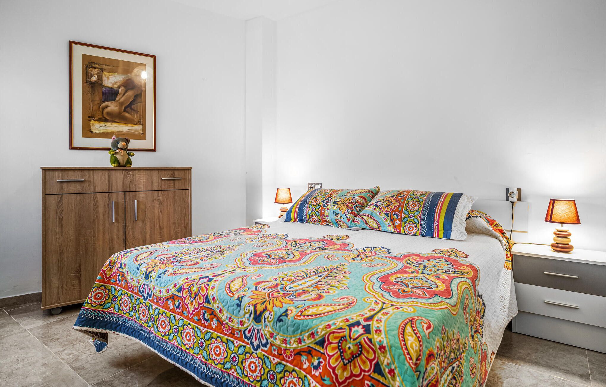 A bedroom with a colorful bedspread, a wooden wardrobe, a framed picture on the wall, and bedside lamps.