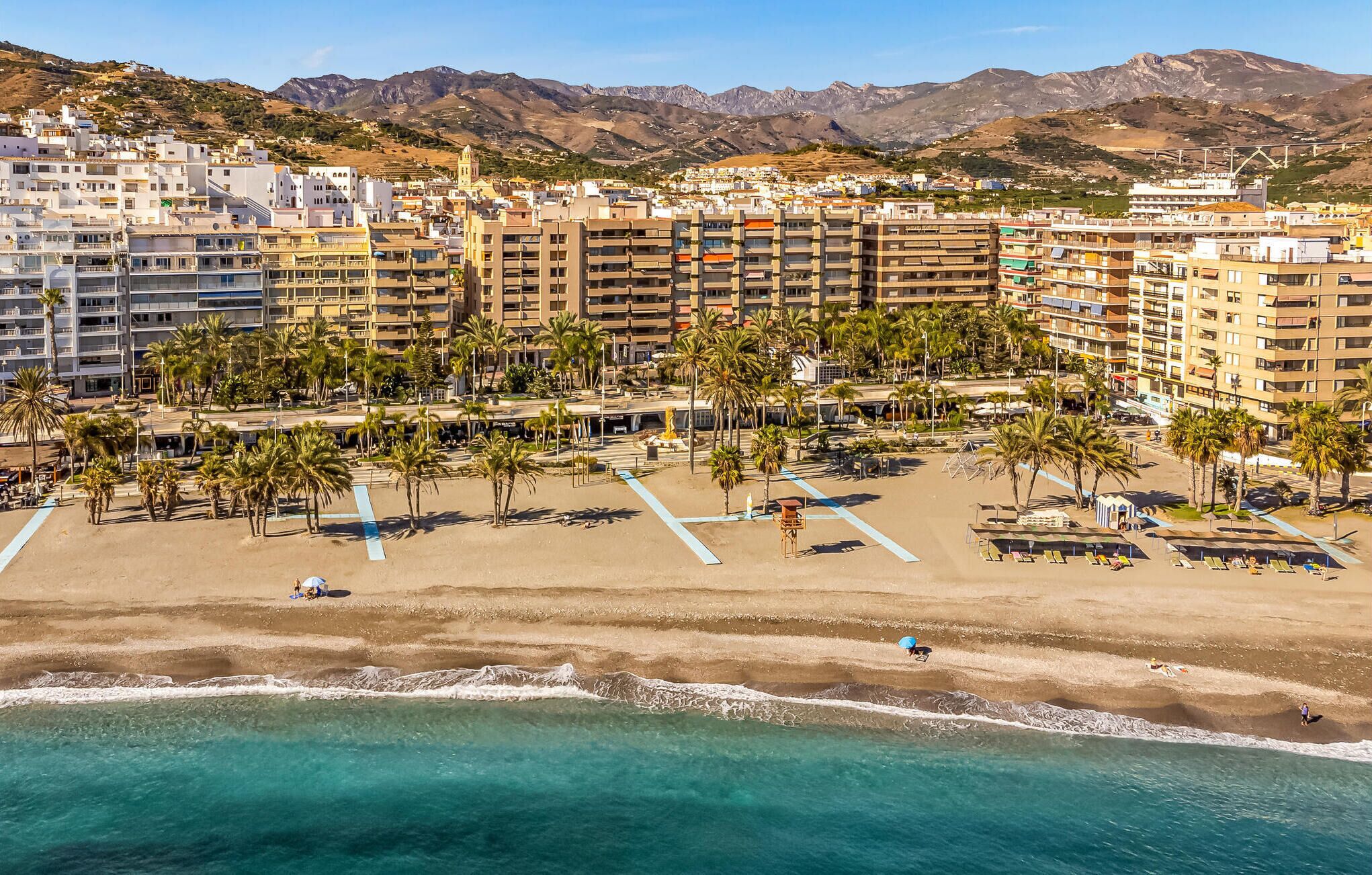 Un complex turístic a la costa amb edificis de diverses plantes, una platja sorrenca amb palmeres i una vista despejada de l'oceà.