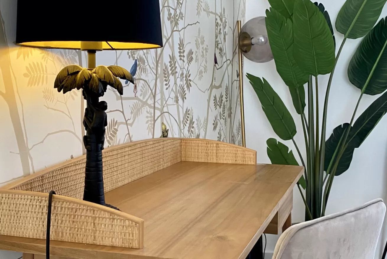 Een hoek van een kamer met een houten tafel, een lamp, een muur met een bladpatroon, en een grote plant.