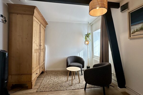 A room with a wooden cabinet, two chairs, a small table, a hanging light fixture, and a window with curtains. - Homebyus, Élégance & Détente en Baie de Somme (Estrébœuf)