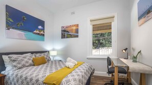 Relax in a serene space with a comfortable chair, desk, and neatly made bed, surrounded by tropical artwork and a view of lush greenery.