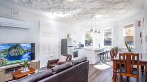Una sala moderna con un televisor de pantalla plana grande, un sofá de cuero marrón, una mesa de comedor de madera con sillas, y una cocina con encimera blanca y taburetes de bar.