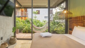 A modern bedroom with a large bed, a hammock, and a view of a lush garden and building outside. - Luxury Condos Tulum - Private Cenote (Tulum)