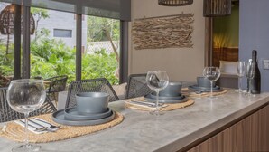 A dining area with a long table set for four, featuring grey plates, bowls, and cutlery. There are two wine glasses on the table, and a bottle of wine is visible on the counter. The room has a modern design with wicker chairs and a view of greenery outside. - Luxury Condos Tulum - Private Cenote (Tulum)