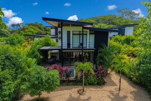 A modern two-story house with a balcony, surrounded by lush greenery and a swimming pool.