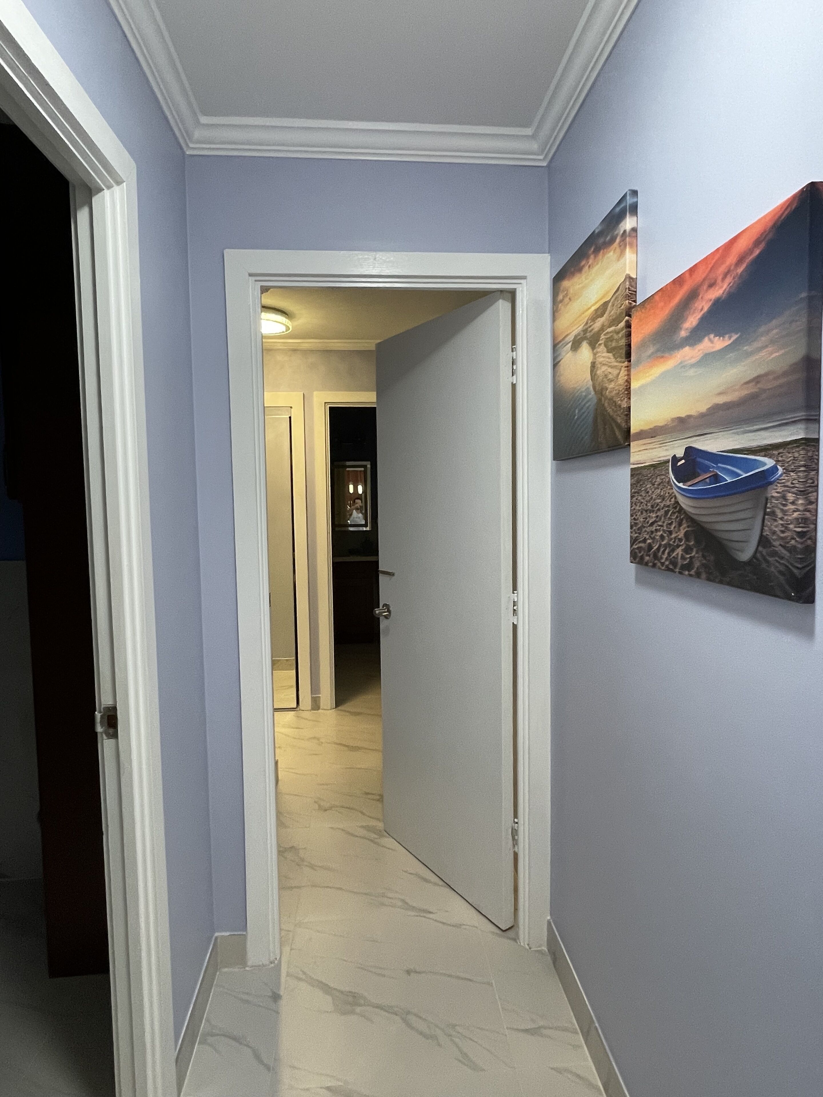 A hallway with a door leading to a room, a painting of a boat on a beach, and a tiled floor.