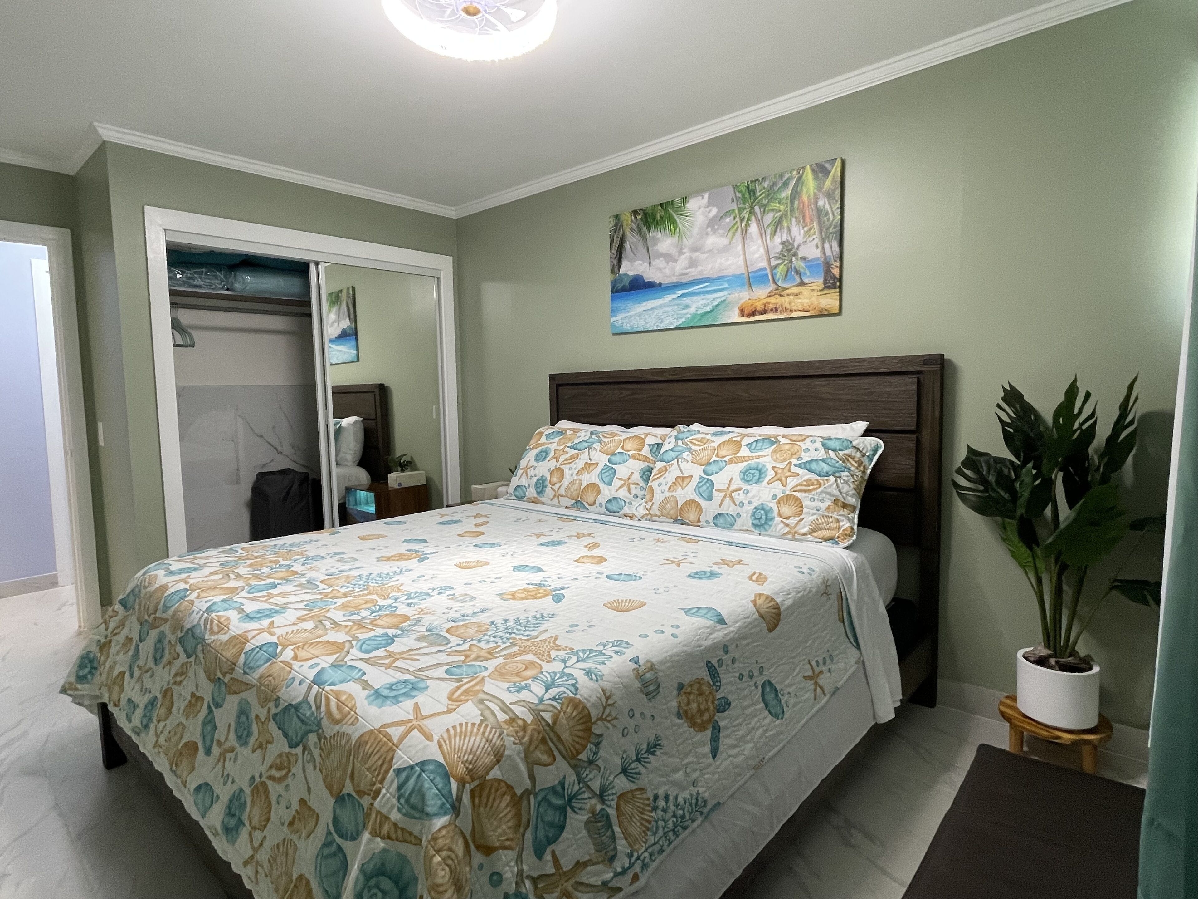 A bedroom with a bed, a nightstand, a potted plant, and a painting of a beach scene.