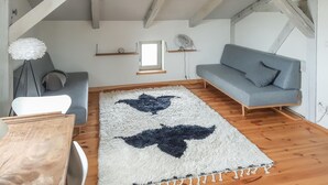 Een gezellige kamer met een grijze bank, een wit tapijt met een sterrenpatroon, houten balken aan het plafond en een klein raam.