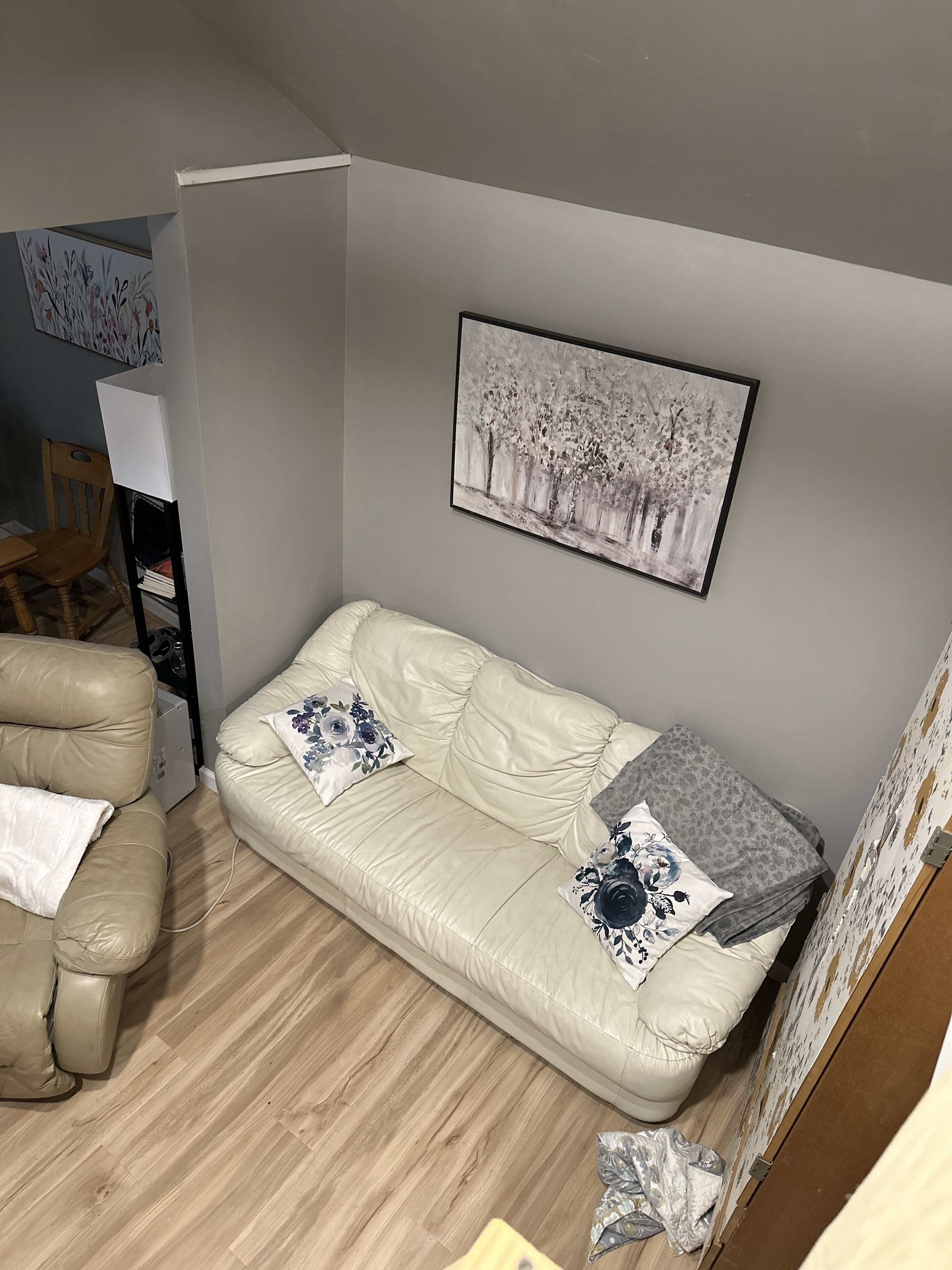 A living room with a beige leather sofa, two pillows, a framed painting, a wooden chair, and a small table.