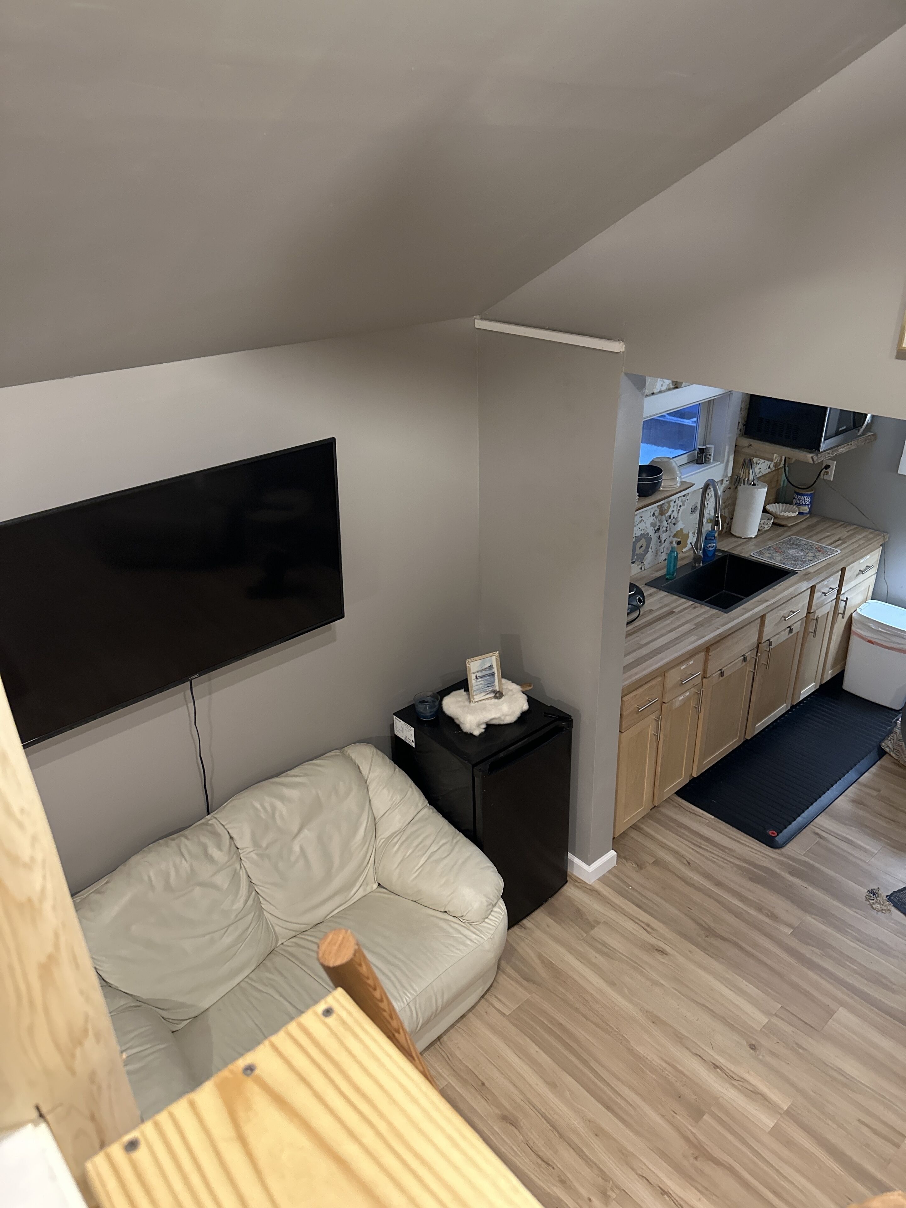 Un espace de vie compact comprenant un canapé, un téléviseur fixé au mur, une petite table d’appoint noire avec un cadre photo, et un coin cuisine avec un évier et un plan de travail.