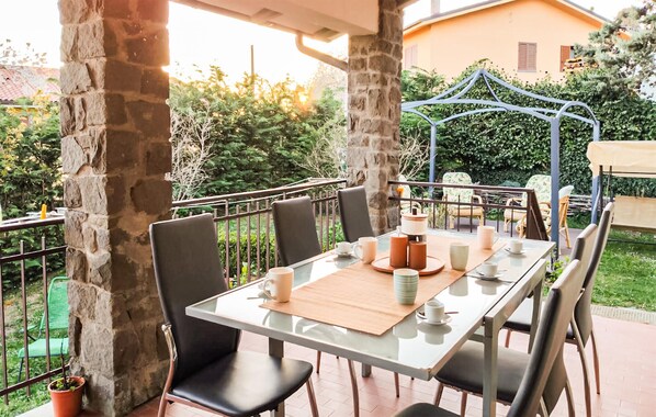 Eine Terrasse mit Tisch für Zwei, umgeben von Steinsäulen, einer Pergola und Blick auf ein Haus und viel Grün.