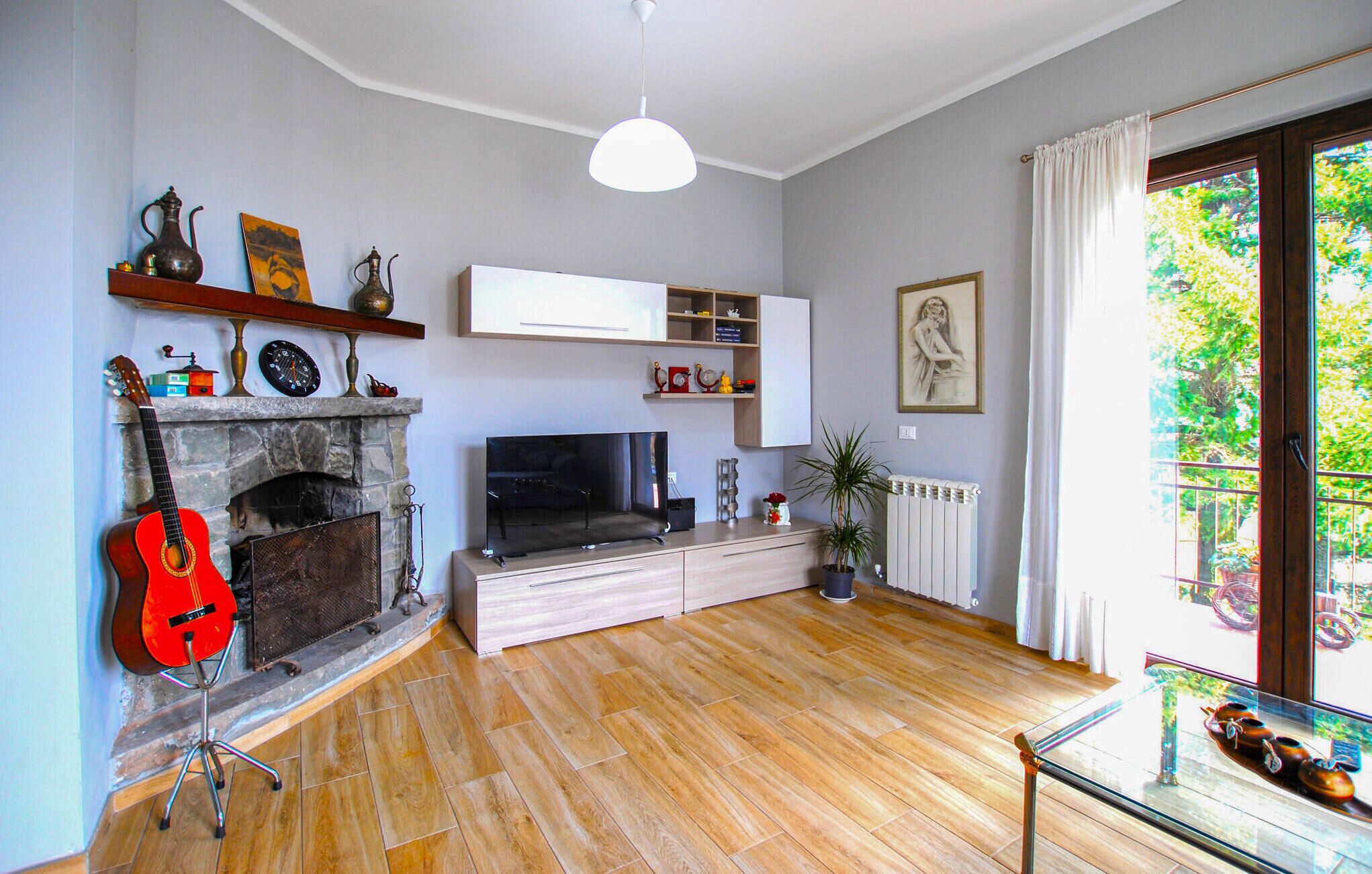 A living room with a fireplace, a TV, wooden flooring, a glass coffee table, and a view of greenery outside.