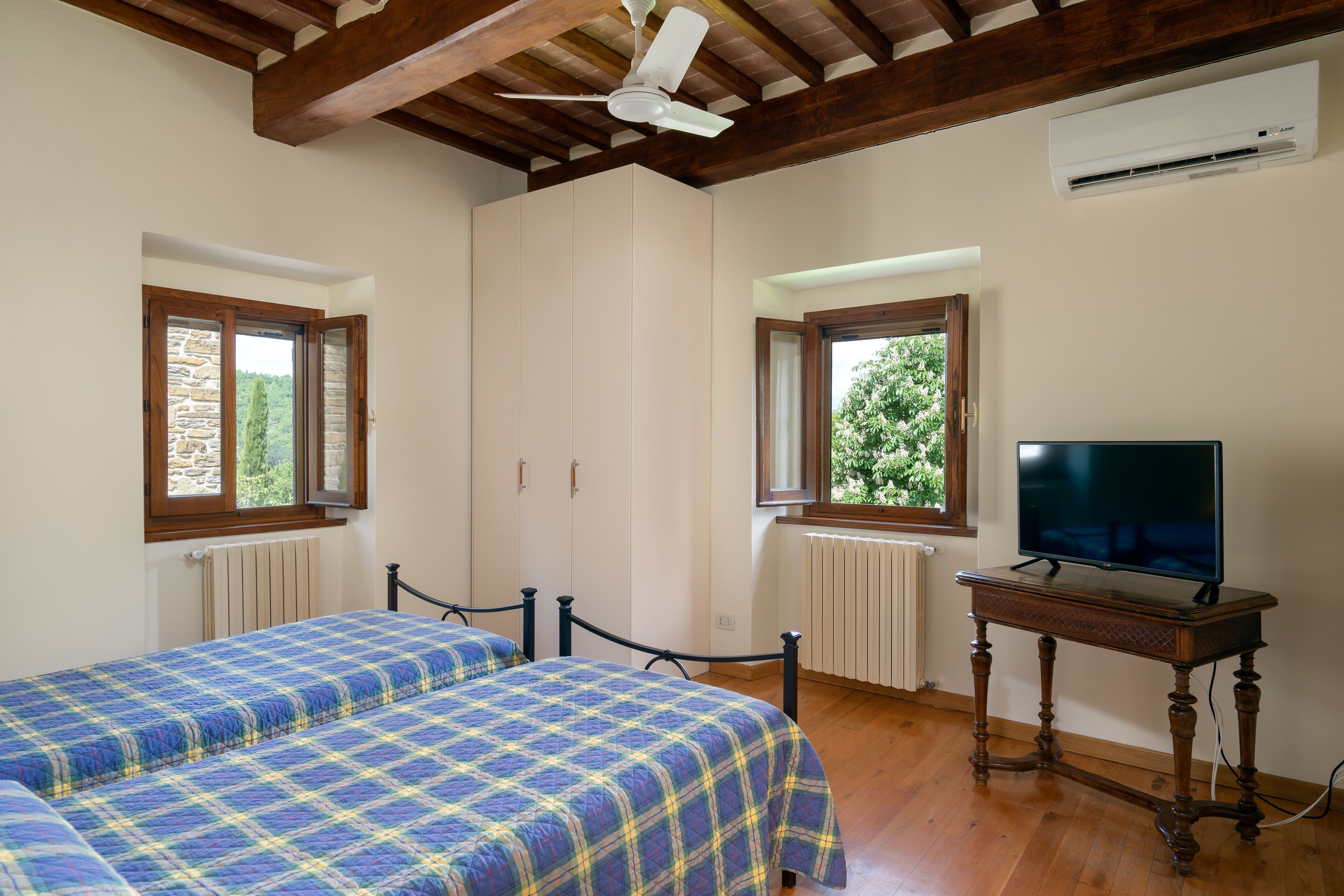 Un dormitorio con dos camas, ventilador de techo, vigas de madera, un televisor sobre una mesa de madera y ventanas con vistas a zonas verdes.