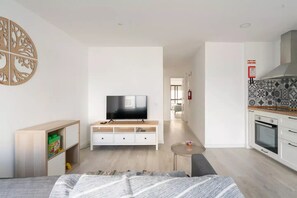 Ein modernes, helles Zimmer mit einem Flachbildfernseher auf einem Holztv-Schrank, einem kleinen runden Tisch, einer Couch und einer Küchenzeile mit gefliestem Fliesenspiegel und einem Herd.