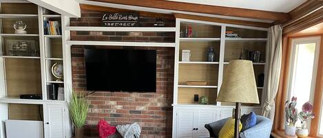 Uma sala de estar acolhedora com um sofá de couro, uma poltrona azul e uma mesa de centro de madeira. Uma TV de tela plana está fixada em uma parede de tijolos, e há prateleiras com livros e objetos decorativos.
