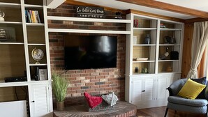 A living room with a brick fireplace, a flat-screen TV mounted above it, and a bookshelf filled with books and decorative items.