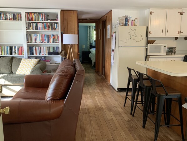 Ein Wohnzimmer mit braunem Ledersofa, einer Bücherwand voller Bücher, einem Holzschrank und einer Küchenzeile mit einem weißen Kühlschrank und schwarzen Barhocker.