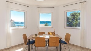 Un espace repas comprenant une table et des chaises, de grandes fenêtres offrant une vue sur la mer et les collines, ainsi qu’un luminaire au plafond.