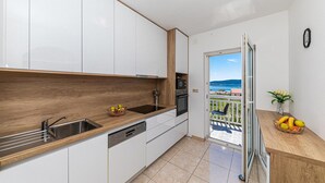 Une cuisine moderne avec des meubles blancs, des plans de travail en bois et une vue sur la mer, accessible par une porte-fenêtre donnant sur un balcon.