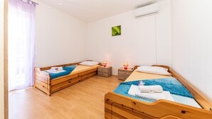 Une chambre avec deux lits en bois, une fenêtre ornée de rideaux violets, un petit climatiseur fixé au mur et un tableau représentant une feuille verte accroché au mur.