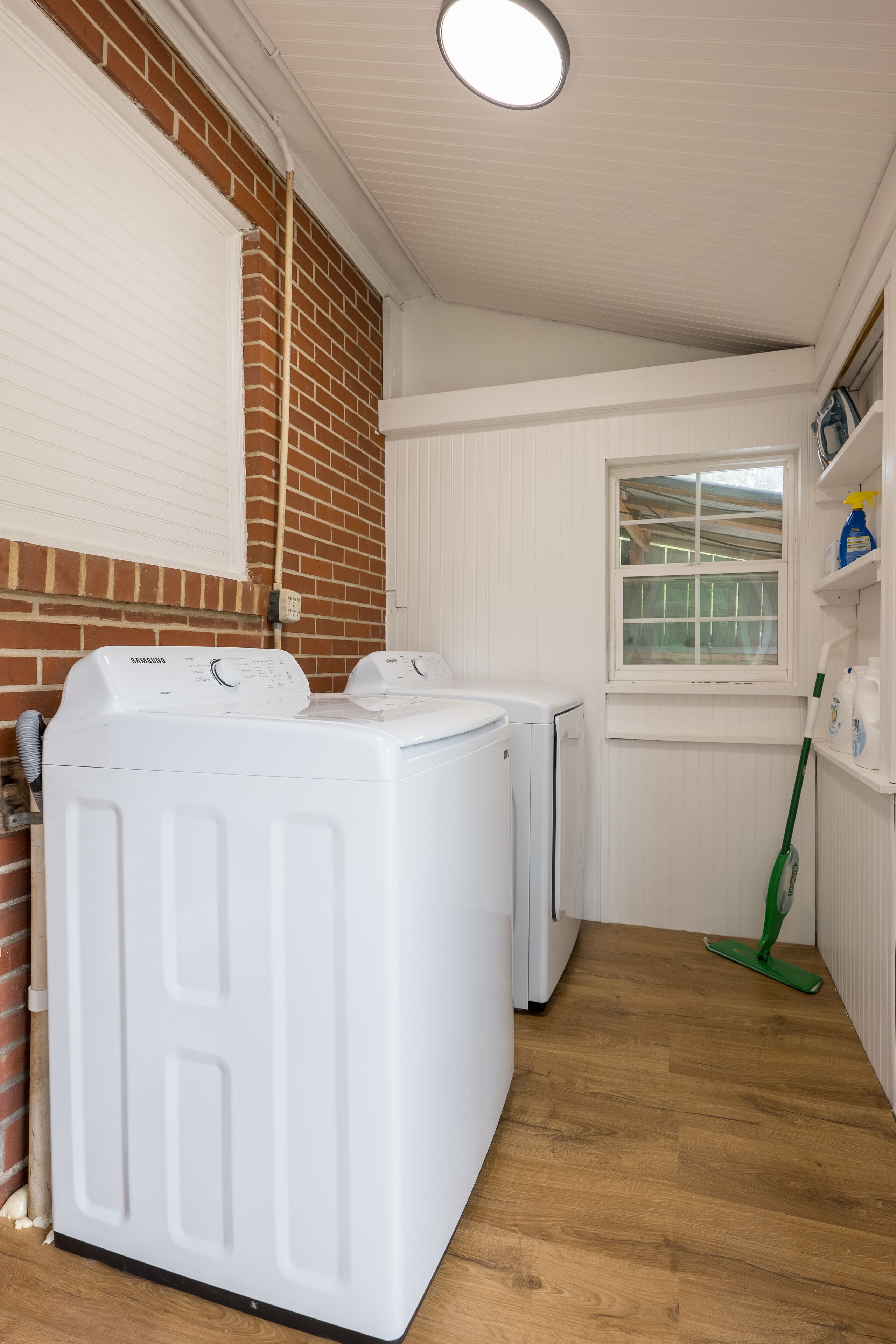 A laundry room with two white front-loading washing machines, a window, and a green mop leaning against the wall.