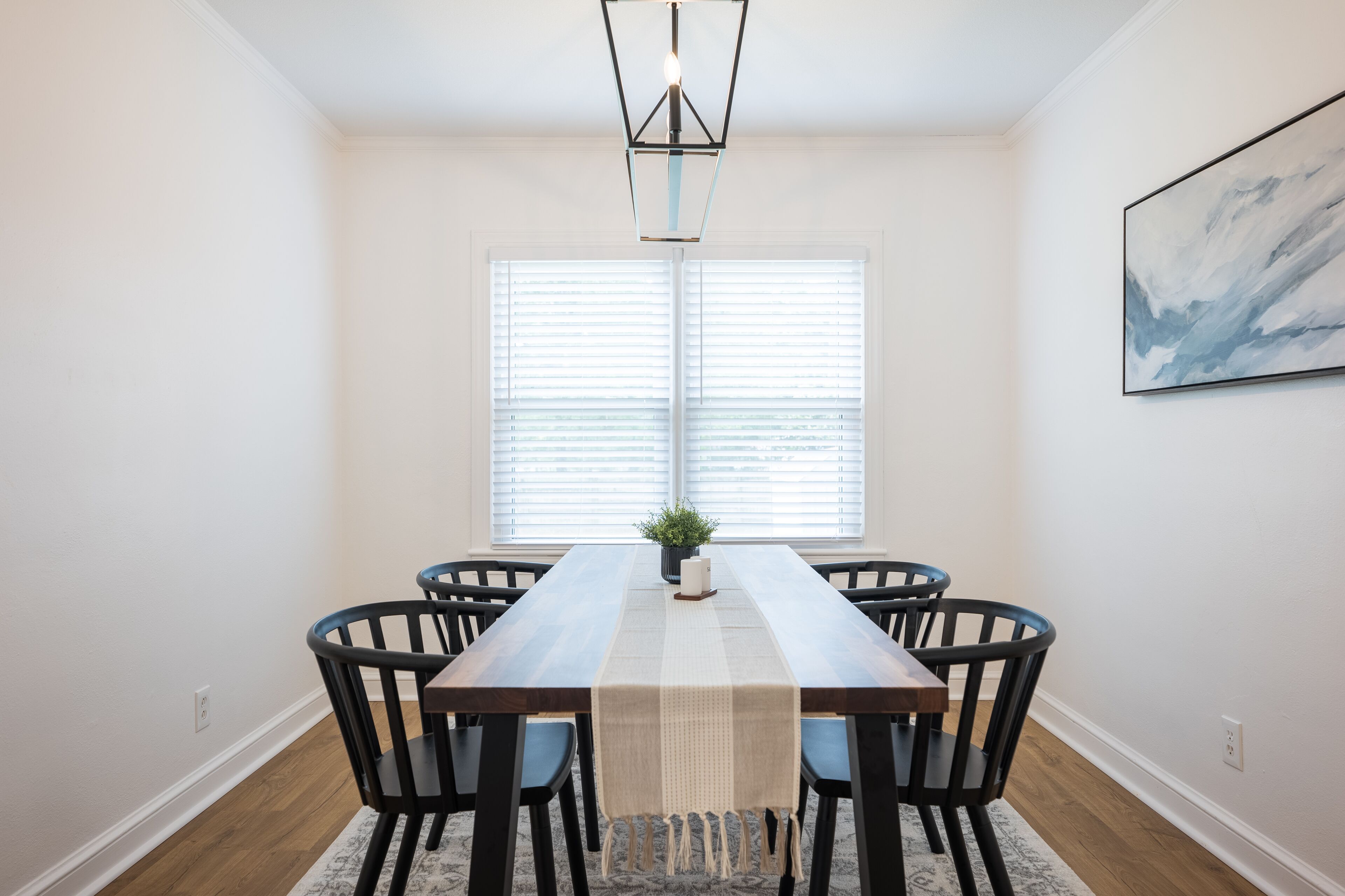 A dining area with a long wooden table, six chairs, a central plant, and a hanging light fixture.