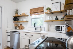A modern kitchen with white cabinetry, a dishwasher, a sink, and a microwave. There are wooden shelves above the counter holding various kitchen items. - The 12th Street Retreat, King Beds, Pets Allowed (Tifton)