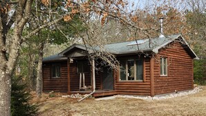 Eine rustikale Holzjagdhaus mit Veranda und Schornstein, umgeben von Bäumen und einer trockenen Landschaft.