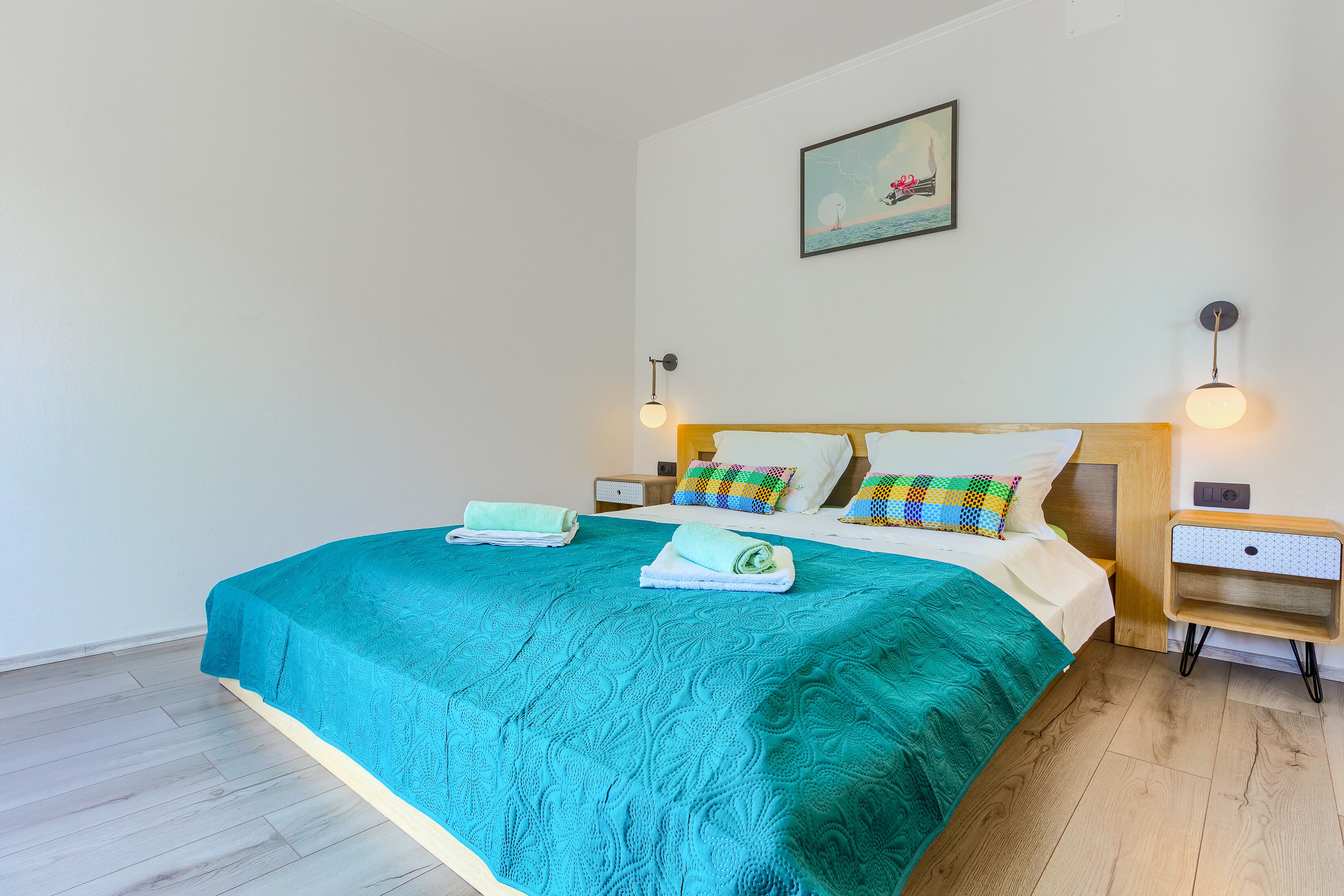 A neatly made bed with a teal bedspread, two pillows, and a matching blanket folded at the foot of the bed. There are two bedside tables with lamps, a wall-mounted artwork, and a wooden floor.