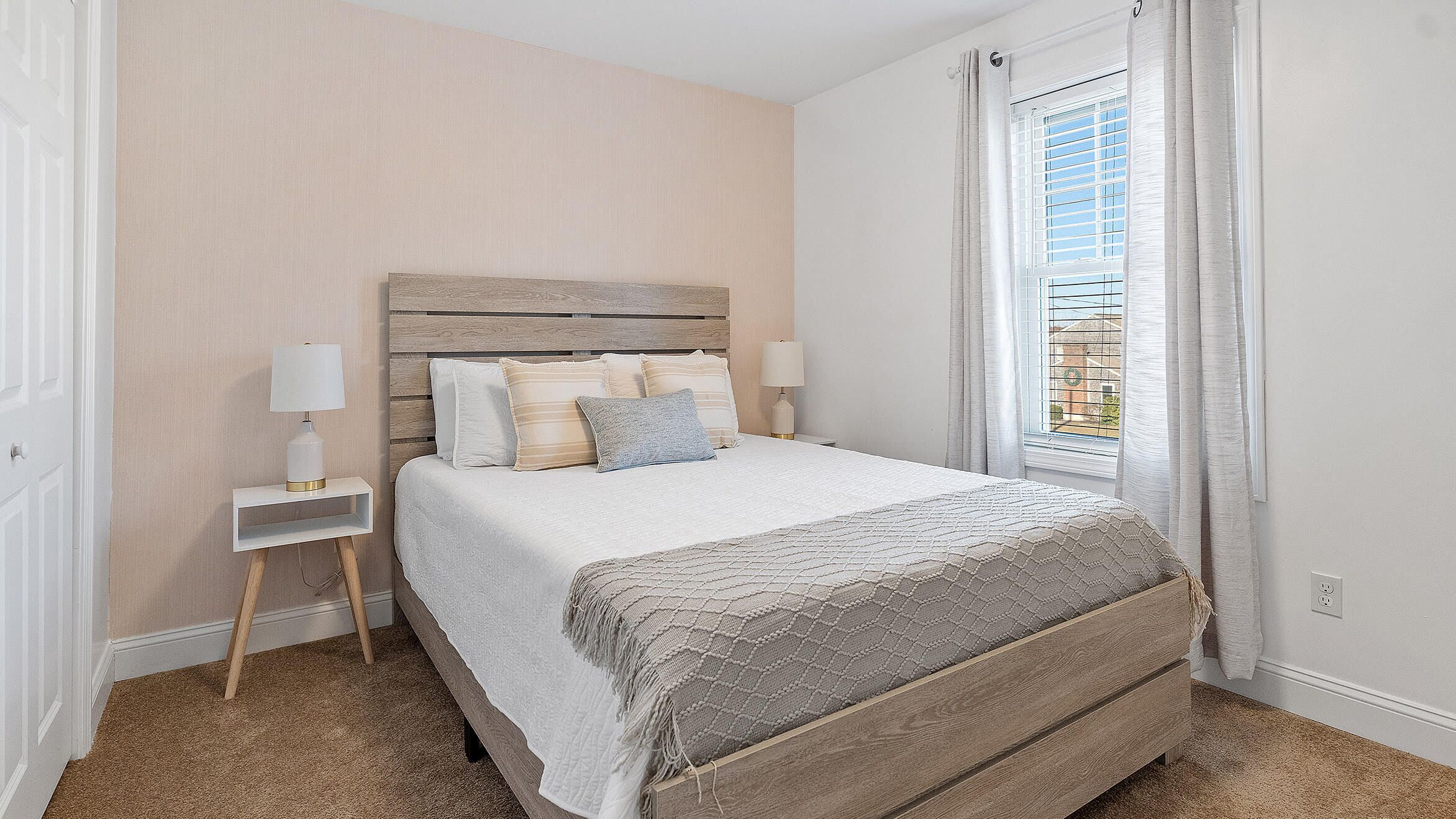 Een slaapkamer met een houten hoofdeinde, een bed met witte lakens, twee nachtkastjes met lampen en een raam met gordijnen.